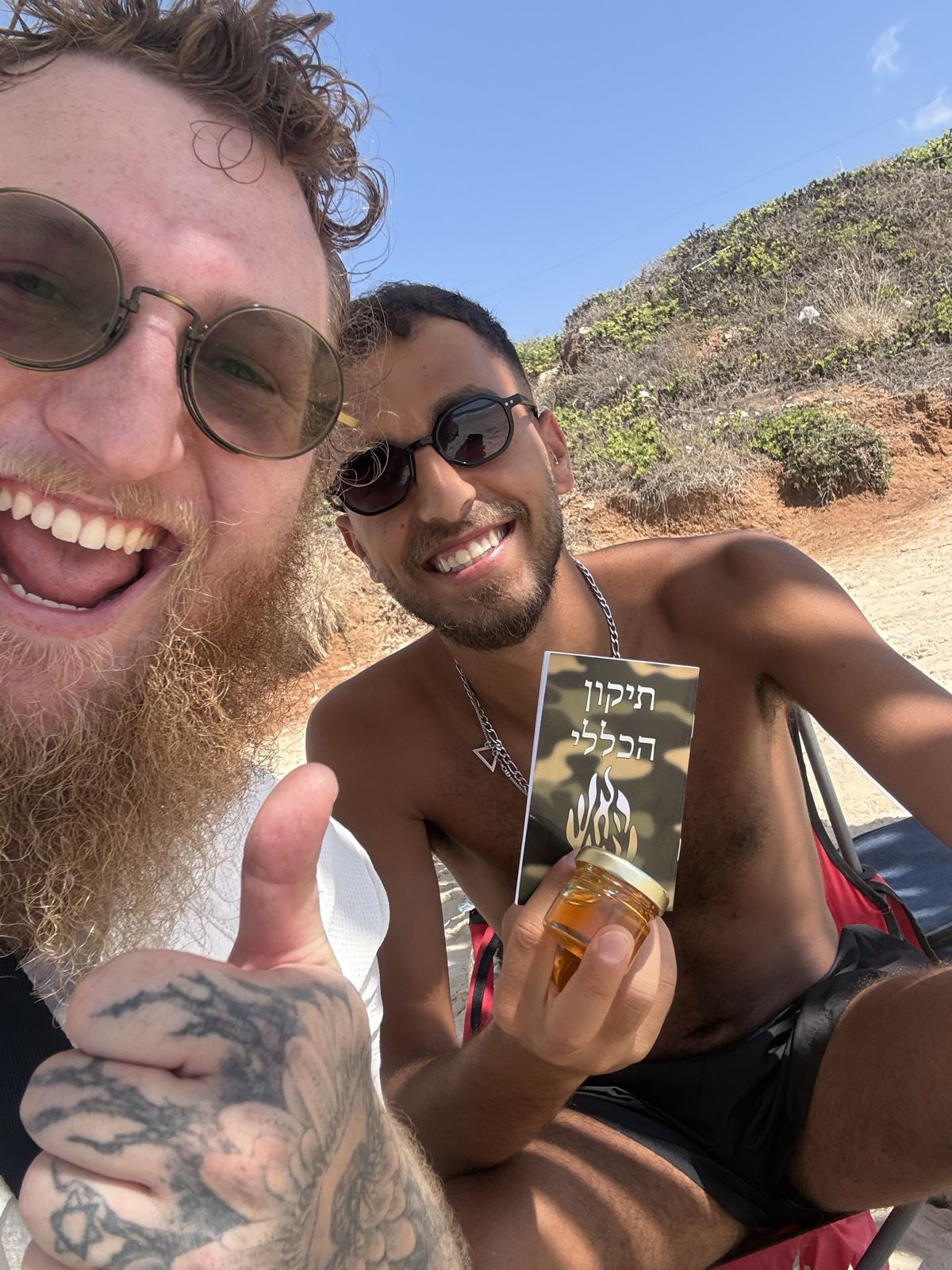 Two men smiling for a selfie on the beach, one holding a small bottle and a card