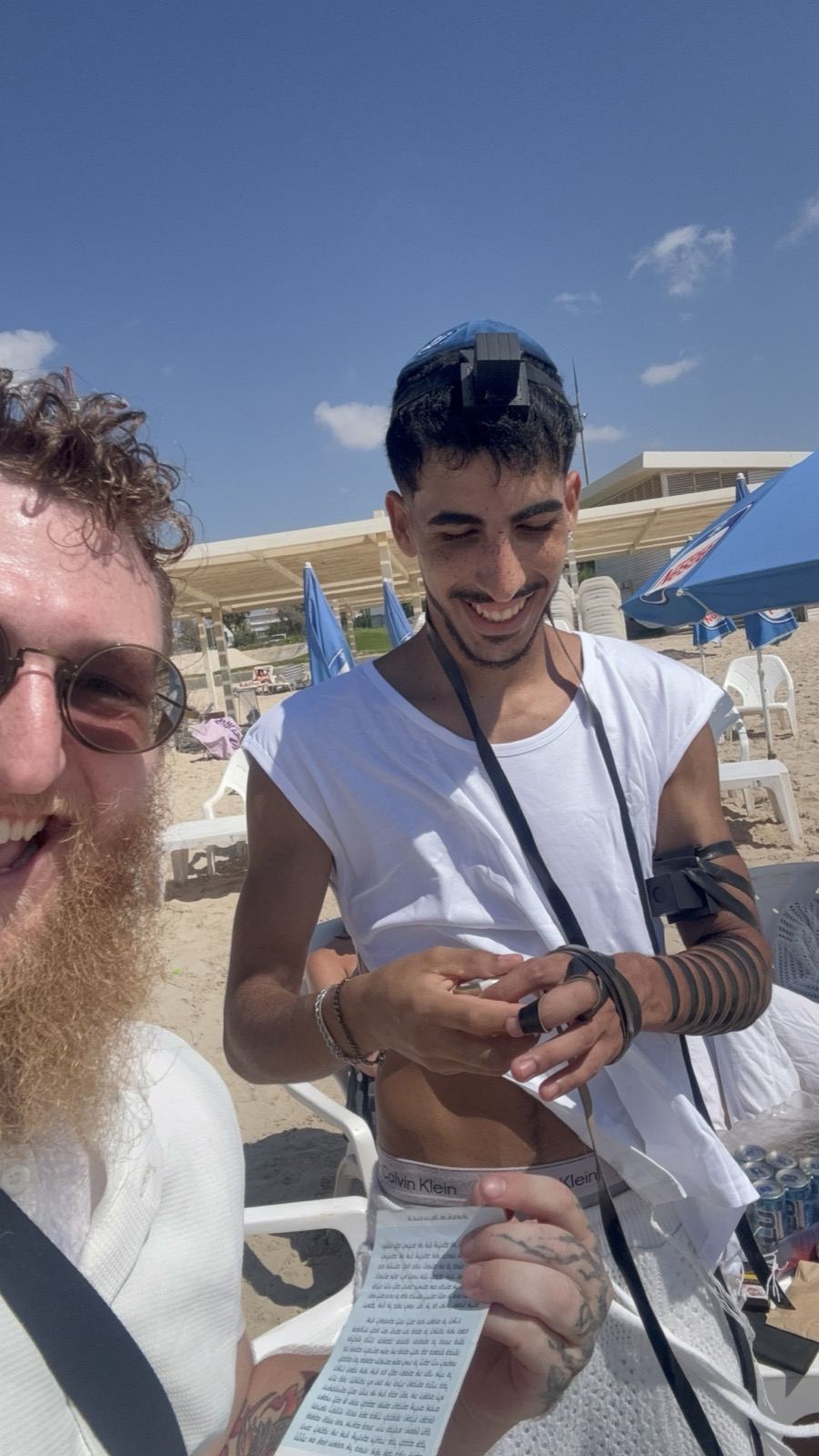 A man wrapping tefillin on the beach while reading from a prayer card