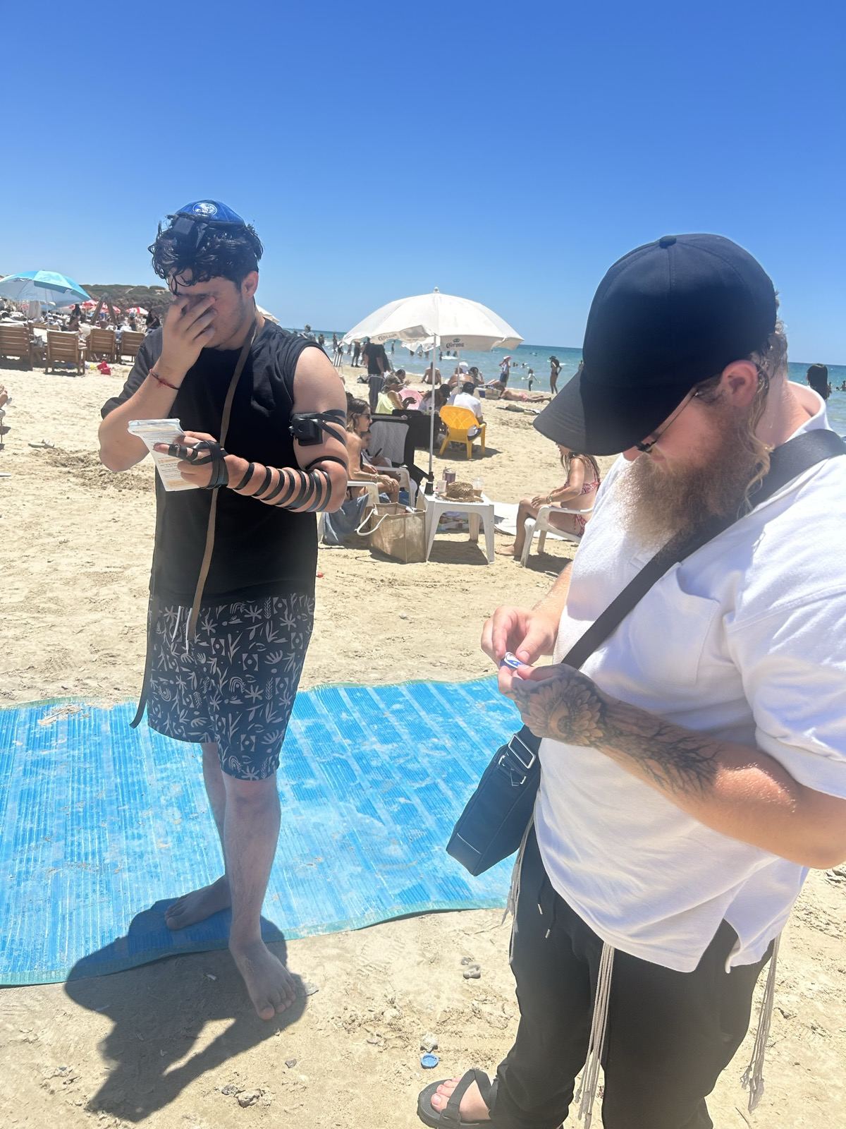 Two men on the beach, one wearing tefillin and reading a prayer card