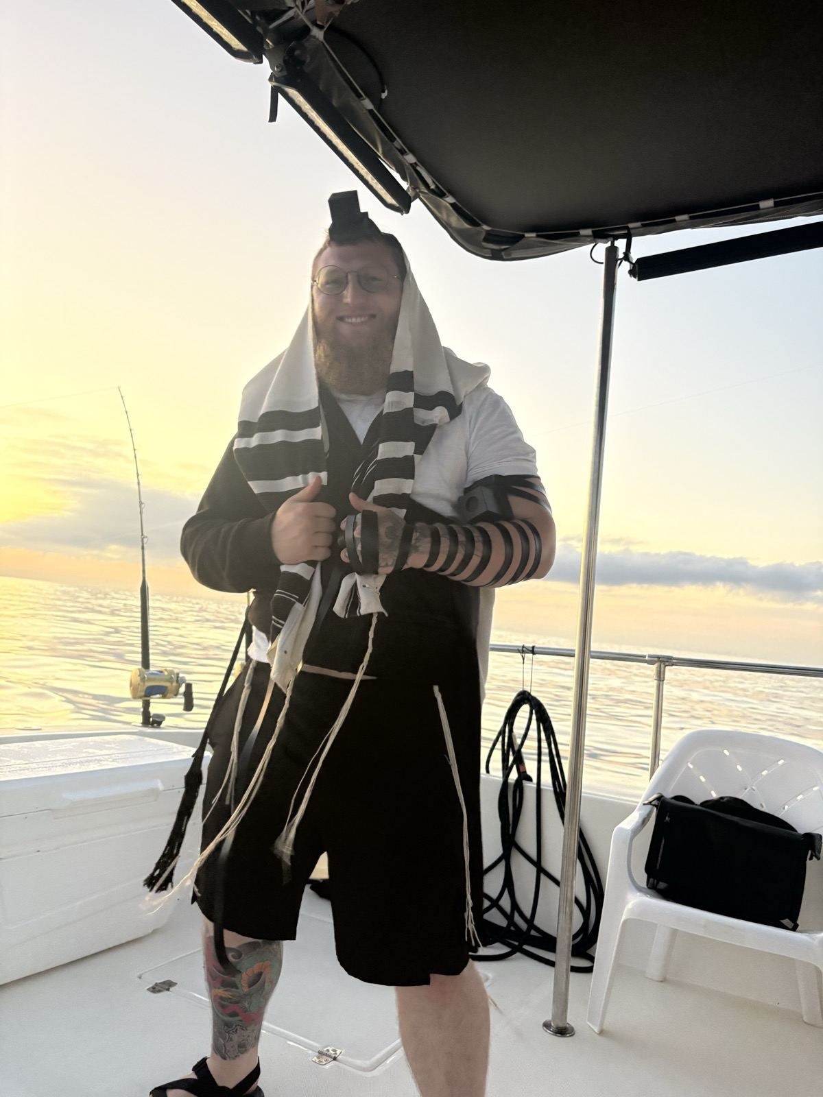 A man wrapped in tefillin and a tallit on a fishing boat at sunset